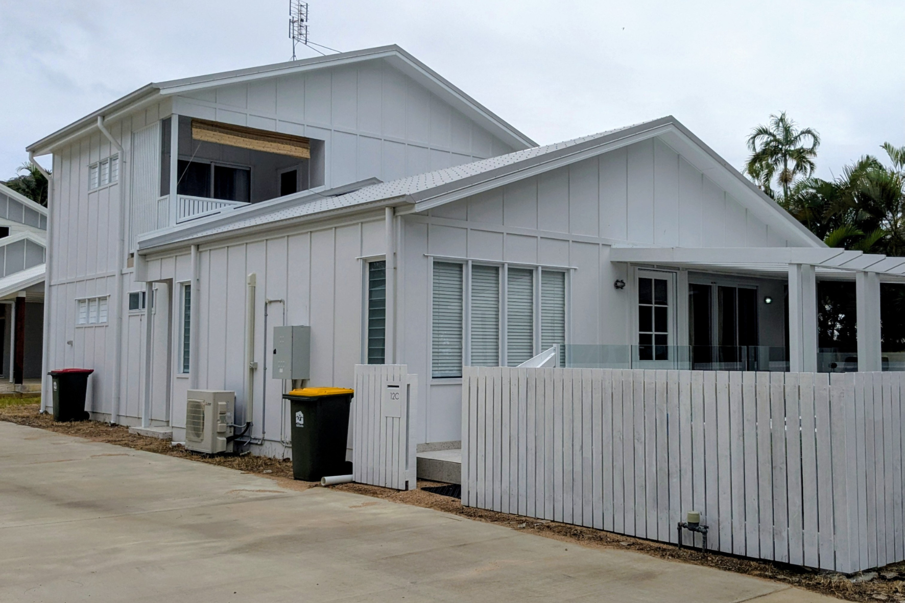 Beach House — completed front elevation from street showing white cladding, upper balcony, pergola and palm trees
