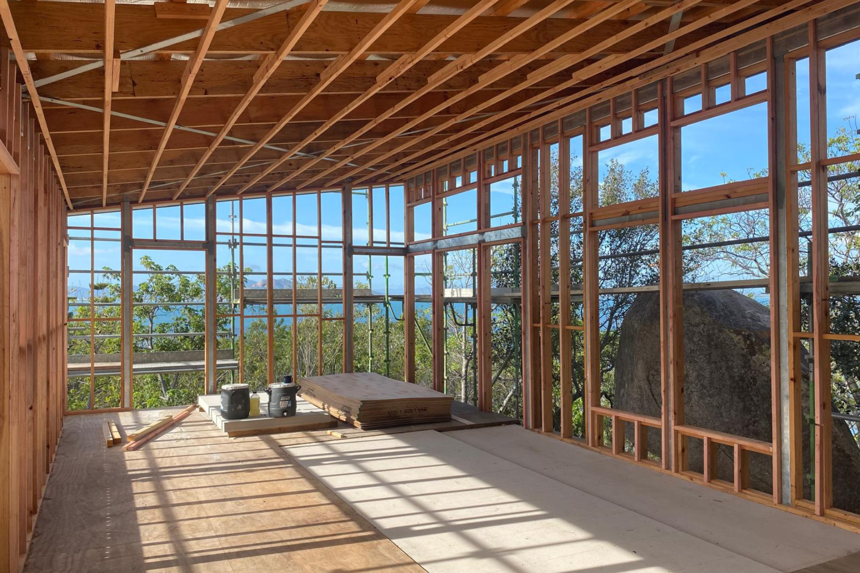Bush Retreat — timber frame interior under construction with full-height glazing openings and water views through the trees