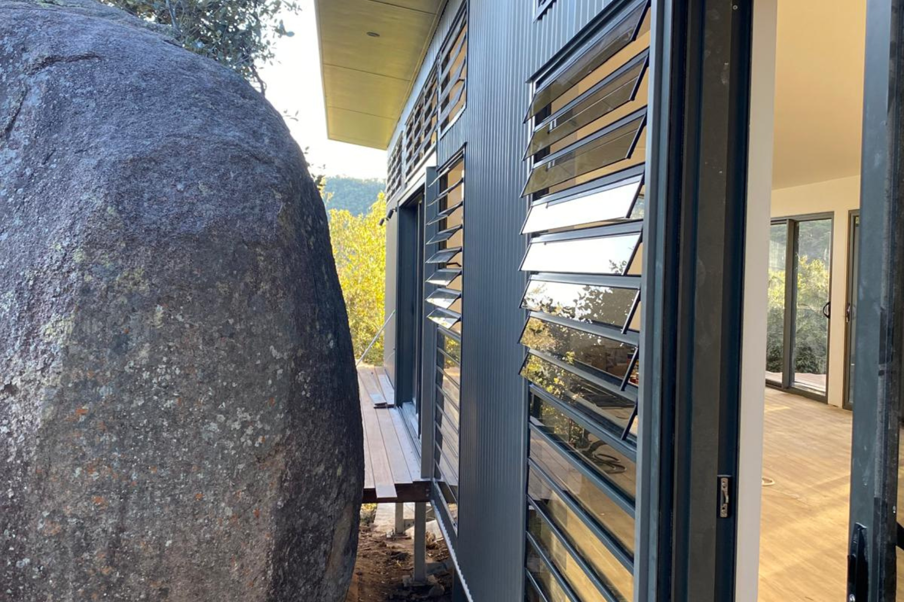 Bush Retreat — close-up of louvre windows and corrugated cladding beside a large granite boulder with timber deck beyond