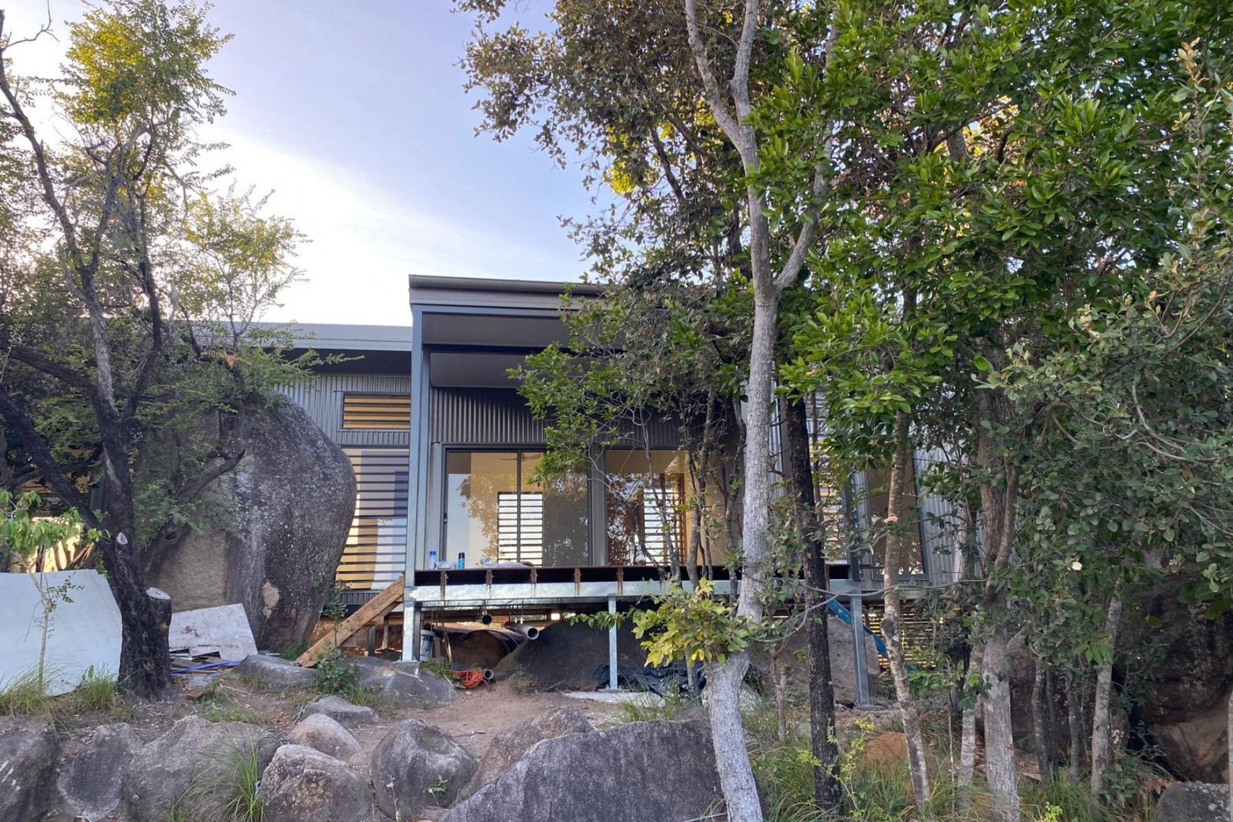 Bush Retreat — exterior view through native trees showing corrugated cladding, louvres and steel subframe under construction