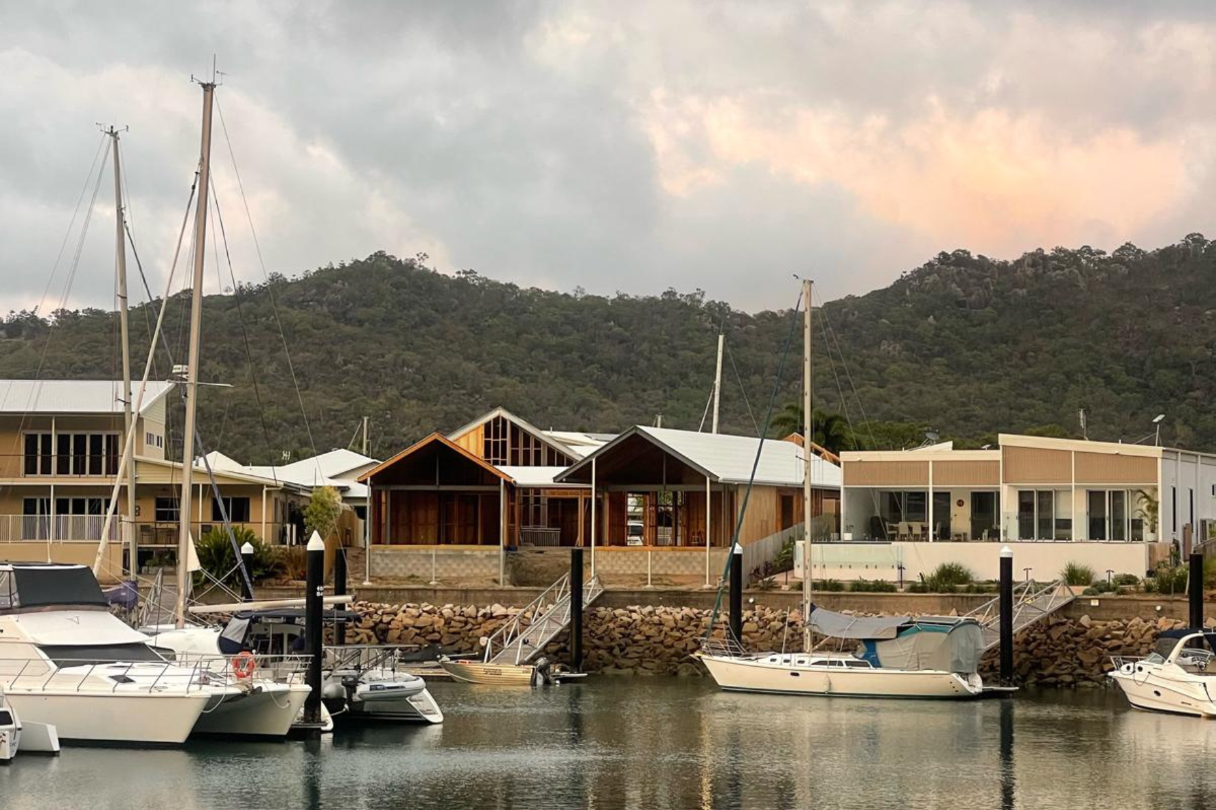 Marina — completed building viewed from the water with sailboats in foreground, timber gable pavilion and mountain backdrop under overcast sky